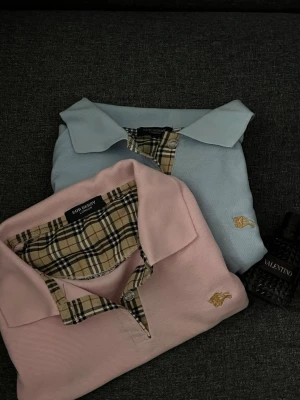 Burberry pikétröja med rutiga detaljer - Säljer två klassiska Burberry pikétröjor, en i ljusblått och en i ljusrosa. Båda har den ikoniska beige-rutiga kragen och knappslån samt broderad logga i guld på bröstet. Perfekt för dig som gillar stilrena och tidlösa plagg med lyxig känsla.köp båda för 750kr eller en för 400kr!