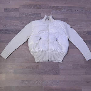 Vit pufferjacka från Moncler - Säljer en riktigt snygg vit pufferjacka från Moncler med stickade ärmar och hög krage. Jackan har två dragkedjeförsedda fickor fram och bred ribbad mudd i midjan och vid ärmsluten. Stor Moncler-logga broderad på ryggen och diskret patch på ärmen.
