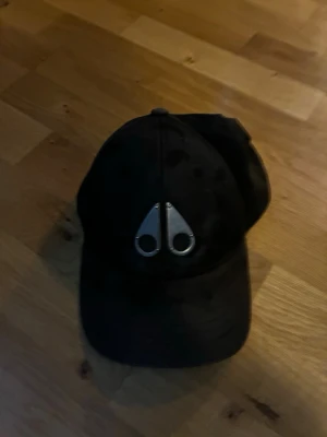 Svart keps Moose Knuckles - Säljer en svart keps från Moose Knuckles med den ikoniska metall-loggan framtill. Kepsen är tillverkad i 100% bomull och har justerbart spänne bak med logodetalj. Perfekt accessoar för en clean och stilren look.