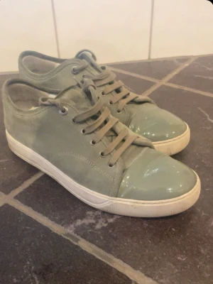 Gröna sneakers med lackad tå från Lanvin - Säljer ett par gröna Lanvin sneakers i mocka med unik blank lackad tå och vita sulor. Skorna har snörning och en stilren, låg profil. Insidan är fodrad i skinn för extra komfort. Perfekta för dig som vill ha något som sticker ut. Extrasnören och boxen ingår hör av er vid fundering eller förslaga!