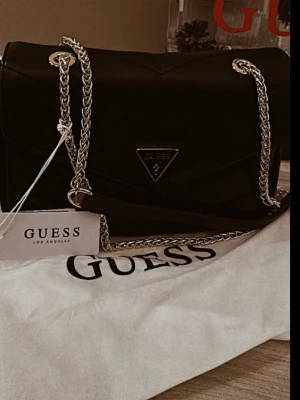 Svart axelväska från Guess - Snygg svart axelväska från Guess med guldfärgad kedjerem och klassisk trekantslogga framtill. 