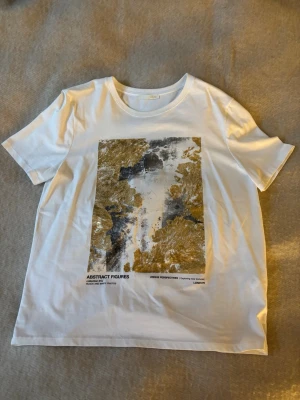 Vit t-shirt med abstrakt tryck från Zara - Säljer en vit t-shirt från Zara med ett stort abstrakt tryck i guld och svart på framsidan i storlek L. T-shirten har rund halsringning och korta ärmar. T-shirten är endast använd ett fåtal gånger och därför i mycket bra skick🤍