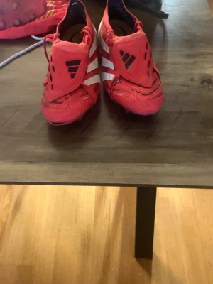 Adidas Predator röda fotbollsskor - Säljer ett par Adidas Predator fotbollsskor i rött med vita och svarta detaljer. Skorna har snörning, låg profil och klassiska tre ränder på sidan. Ovandelen är i syntetmaterial och sulan har dobbar för bra grepp på planen. Perfekta för dig som vill sticka ut på matchen.