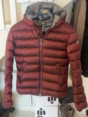 Vinröd pufferjacka från Colmar - Säljer en vinröd pufferjacka från Colmar med huva och två sidofickor. Jackan har en rejäl dragkedja framtill och är quiltad för extra värme. Perfekt för kalla dagar och har en snygg, sportig look. Märkeslogga på ärmen för extra detalj. Den är äkta. Mycket bra skick. 💞💞