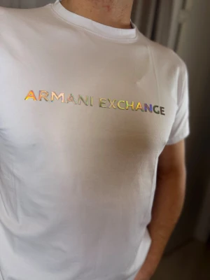 Vit t-shirt från Armani Exchange - Snygg vit t-shirt från Armani Exchange med holografiskt tryck över bröstet. Klassisk rund hals och korta ärmar. Tillverkad i mjuk bomull som känns skön mot huden. Perfekt för dig som vill ha en clean look med en twist.