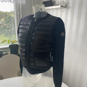 Svart Moncler cardigan dam - Svart cardigan jacka från Moncler med quiltad, glansig framsida och två fickor med knapp. Ärmar och rygg är stickade för en snygg kontrast. Dragkedja framtill och klassisk Moncler-logga på ärmen. Perfekt för dig som vill ha en stilren och exklusiv look.