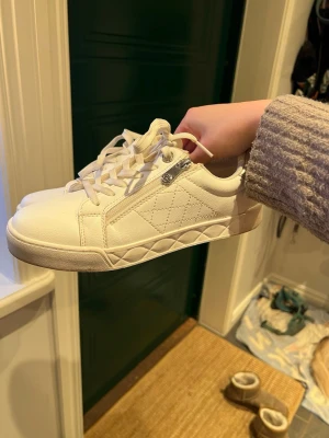 Vita sneakers från Ceci Comfort strl 38 - Säljer ett par vita sneakers från Ceci Comfort i storlek 38. Skorna har quiltade detaljer på sidan, silvriga hälpartier och dragkedja vid snörningen. Snygg och enkel design med platt sula och rund tå. Perfekta till jeans eller kjol!