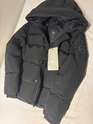 Svart pufferjacka från Canada Goose - Säljer en svart pufferjacka från Canada Goose med huva och diskret logga på ärmen. Jackan har dragkedja och tryckknappar framtill, samt en riktigt schysst, vadderad look som håller dig varm under kalla dagar. Perfekt för dig som vill ha både stil och funktion.