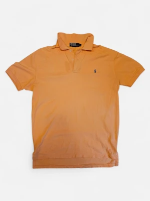 Orange pikétröja från Ralph Lauren - Klassisk orange pikétröja från Polo Ralph Lauren med blå broderad logga på bröstet. Tröjan har korta ärmar, krage och knappar framtill. Tillverkad i mjuk bomull som känns skön mot huden. Perfekt för dig som gillar stilrena och tidlösa plagg.
