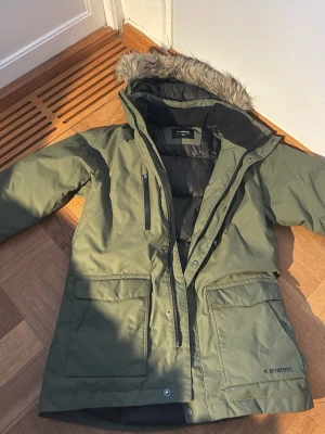 Grön jacka från Everest  - Säljer en grön jacka från Everest med huva och fuskpälsdetalj. Jackan har stora fickor framtill, dragkedja och knappar, samt en bröstficka med dragkedja. Insidan är vadderad och jackan är perfekt för kalla dagar.