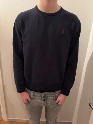Mörkblå sweatshirt från Polo Ralph Lauren - Klassisk mörkblå sweatshirt från Polo Ralph Lauren med röd broderad logga på bröstet. Tröjan har rund halsringning och långa ärmar. Tillverkad i mjuk bomull som är skön att ha på sig. Perfekt till jeans eller shorts.