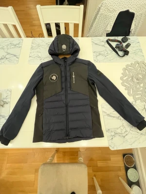 Mörkblå Sail Racing pufferjacka - Säljer en mörkblå pufferjacka från Sail Racing med huva och snygga detaljer som Sail Antarctica Expedition-märke på bröstet. Jackan har dragkedja framtill, två sidofickor med dragkedja och logga på ärmen. Perfekt för kalla dagar och riktigt stilren look.