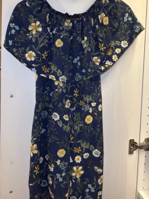 Blommig byxdress  - Säljer en mörkblå byxdress med blommigt mönster i gult, vitt och grönt. Byxdressen har vida ben och en volang upptill som ger en snygg off-shoulder-look. Perfekt för dig som vill sticka ut med färg och mönster. Säljer då den är för lång för mig, passar mer till 165-170 cm i längd. 