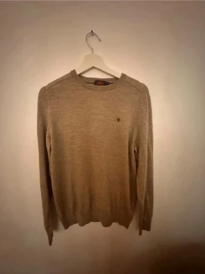 Beige merinoulltröja från Morris - Säljer en stilren beige tröja från Morris i 100% merinoull. Tröjan har rund halsringning, diskret logga broderad på bröstet och långa ärmar. Perfekt för dig som gillar klassisk och enkel stil.