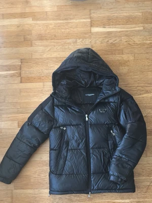 Svart pufferjacka Alessandro Milano S - Säljer min svarta pufferjacka från Alessandro Milano i storlek S. Jackan har huva, dragkedja framtill, två snedställda fickor med dragkedja och snörning i både huva och midjan. Materialet är glansigt och quiltat, perfekt för kalla dagar. Den är inte använd så mycket och är i väldigt bra skick.