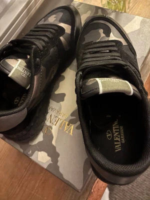 Valentino Garavani camo sneakers - Säljer ett par Valentino Garavani sneakers i svart och grått med camouflagemönster. Skorna har snörning, rund tå och coola nitar på sulan. Materialmix av mocka och skinn ger en lyxig känsla. Perfekta för dig som vill sticka ut med exklusiva detaljer.