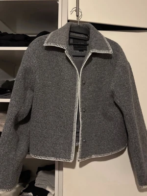 Grå teddyjacka med kontrastsömmar - Säljer en grå teddyjacka med vita kontrastsömmar längs krage, dragkedja och nederkant. Jackan har en boxig passform, klassisk krage och stängs med tryckknappar. Perfekt för dig som gillar en stilren men ändå unik look.
