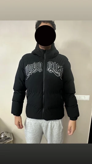 Svart pufferjacka från Hoodrich M - Svart pufferjacka från Hoodrich med stor vit logga på bröstet. Jackan har huva, dragkedja framtill och elastiska muddar vid ärmslut. Materialet är syntet och jackan har en loose passform som ger en chill vibe. Perfekt för kalla dagar när du vill se fresh ut.