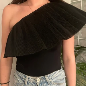 Svart oneshoulder topp med volang - Säljer en svart oneshoulder topp som är en body! med stor plisserad volang som går över axeln och bröstet. 
