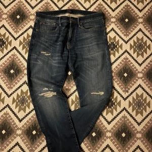 Blå jeans från Polo Ralph Lauren - Snygga blå jeans från Polo Ralph Lauren med slitna detaljer och klassisk femficksdesign. Jeansen har en rak passform och är tillverkade i robust denim med coola slitningar på låren och knäna. Perfekt för dig som gillar en avslappnad och trendig look.
