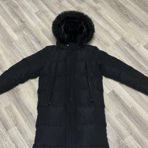 Svart lång pufferjacka Supply & Demand - Lång svart pufferjacka från Supply & Demand med huva och fuskpäls. Jackan har två snedställda dragkedjefickor framtill och en stor logga på ärmen. Insidan av huvan är fodrad med mjuk pälsimitation för extra värme.