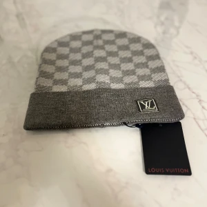 Grå rutig mössa från Louis Vuitton - Snygg grå mössa från Louis Vuitton med klassiskt rutigt mönster och vikt kant. Mössan har LV-logga framtill och är tillverkad i mjukt stickat material. Perfekt accessoar för att lyfta din vinterstil.