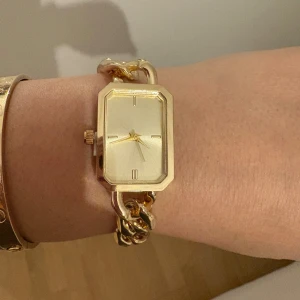 Guldig fyrkantig länkad klocka - Snygg klocka med rektangulärt ur i guldton och kedjelänkad armbandsdesign. Urtavlan är stilren och minimalistisk utan siffror, med guldiga visare och detaljer. Armbandet har breda länkar som ger en cool och modern vibe. Perfekt accessoar för att lyfta din outfit.