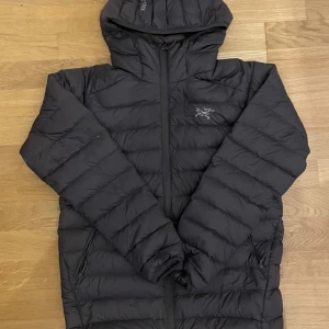Grå dunjacka från Arc'teryx med huva - Säljer en svart dunjacka från Arc'teryx med huva och dragkedja framtill. Jackan har ett stilrent, puffigt utseende och är perfekt för kalla dagar. Storlek M men passar också väldigt bra som storlek S