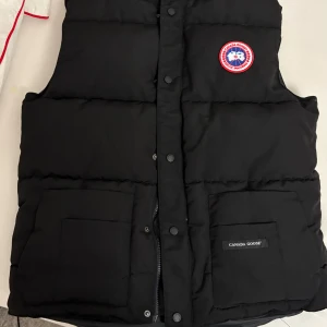 Svart dunväst från Canada Goose - Svart dunväst från Canada Goose med klassisk logotyp på bröstet. Västen har två stora fickor framtill, knappar och dragkedja. Den är quiltad och har en stilren, enkel design som funkar till många olika looks.