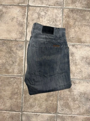 Nudie jeans  - Säljer ett par jeans från Nudie jeans i grå tvätt med snygga slitningar och hål vid knäna. Modellen är slim och har klassisk femficksdesign. Jeansen är i stretchigt denim och passar dig som gillar en smalare siluett med lite edge.Obs modellappen ej kvar. Midja 35cm, längd 103cm. JAG SKICKAR EJ FLER BILDER NÄR JAG BÄR JEANSEN! för storleksguide hänvisar jag till måtten och för bättre inblick i passformen rekommenderar jag att googla på modellnamnet. kolla måtten noggrant :) 45