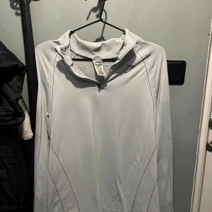 Ljusgrå Gymshark half zip tröja - Snygg ljusgrå långärmad stretchigtröja från Gymshark med half zip och sportig design. Tröjan har diskreta mörka sömmar och Gymshark-logga på bröstet. Perfekt för träning eller chill, tillverkad i mjuk polyester som andas.