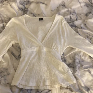 Vit omlottopp med knytband - Säljer en vit omlottopp från Monki med långa ärmar och v-ringad hals. Toppen har snygga knytband i midjan och är tillverkad i ett lätt, krinklat tyg som ger en cool struktur. Perfekt att styla med jeans eller kjol för en fräsch look.