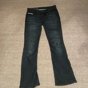 Mörkblåa bootcut jeans från Diesel - Snygga mörkblåa jeans från Diesel med bootcut passform och klassisk femficksdesign. Byxorna har diskreta slitningar och Diesel Industry-detaljer på fickan och bakre läderpatch. Perfekta för dig som gillar en cool och tidlös look. Frakt tillkommer!