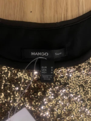 Guldig paljettklänning från Mango - Supersnygg kort klänning från Mango, täckt med glittrande guldiga paljetter. Modellen är figurnära med rund hals och korta ärmar, perfekt för att sticka ut på festen. Materialet är polyester och klänningen har en lyxig känsla med sin glittriga yta.