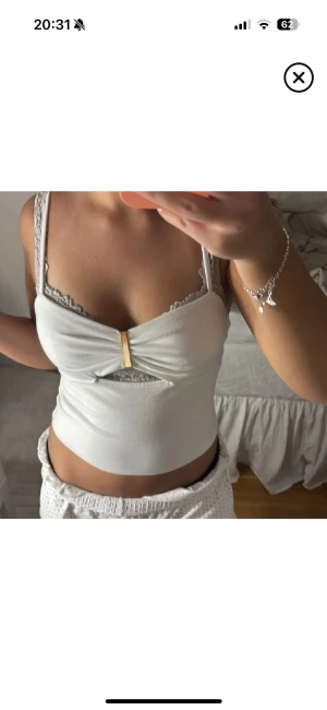 Vit croppad linnetopp med gulddetalj - Säljer en vit croppad linnetopp med smala axelband och snygg guldfärgad detalj framtill. Toppen har en cut-out under bysten och spetskant vid urringningen som ger en extra touch. Perfekt för dig som gillar stilrena och trendiga plagg.