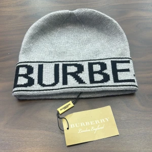 Grå stickad Burberry mössa - Snygg grå mössa från Burberry med bred svart logotyp runt kanten. Stickad i mjukt material som håller dig varm och ger en clean look. Perfekt för dig som vill ha en stilren accessoar med lyxig känsla och tydlig branding.
