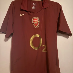 Arsenal Highbury 2005/06 Nike tröja - Säljer en ikonisk Arsenal Highbury 1913-2006 fotbollströja från Nike. Tröjan är vinröd med guldfärgade detaljer, klubbmärke och O2-logga på bröstet. Klassisk krage och korta ärmar. Perfekt för dig som älskar retro och Arsenal.