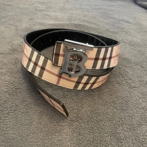 Burberry bälte med rutig design - Snyggt Burberry bälte med klassiskt rutig mönster i beige, svart och rött på ena sidan och svart rutig design på andra. Bältet har en stor silvrig metallspänne med 'B'-logga och är gjort i skinn. Perfekt accessoar för att lyfta din outfit.
