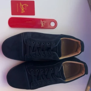 Mocka sneakers från Louboutin - Säljer ett par mörkblåa sneakers från Louboutin i mjuk mocka med klassisk röd sula och snygg signatur på hälen. Skorna har rund tå, platt sula och snörning framtill. Insidan är beige och ger en lyxig känsla. Perfekt för dig som gillar exklusiva detaljer.