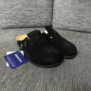 Birkenstock Boston svarta mockasandaler - Birkenstock Boston sandaler i svart mocka med justerbart spänne och korksula. Modellen har en sluten ovandel och platt sula med mönstrad undersida för extra grepp. Perfekt för dig som gillar klassisk och chill stil.