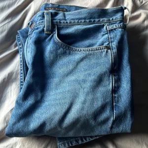 Nudie jeans Gritty jackson - Säljer ett par klassiska blå jeans från Nudie Jeans med snygga orangea sömmar och logga på bakfickan. Jeansen har fem fickor, normal passform och är tillverkade i slitstarkt bomullsjeansmaterial. Perfekt för dig som gillar tidlös stil och kvalitetsjeans.