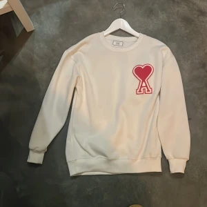 Vit sweatshirt från Ami med rött hjärta - Säljer en clean vit sweatshirt från Ami Alexandre Mattiussi med ett stort rött hjärta och bokstaven A broderat på bröstet. Tröjan har rund hals, ribbade muddar och lång ärm. Perfekt för dig som gillar stilrena detaljer och vill sticka ut lite. Pris kan diskuteras.