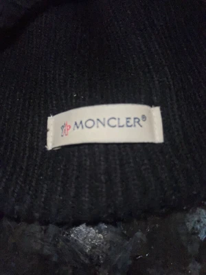 Svart Moncler mössa i ribbstickad ull - Säljer en svart ribbstickad mössa från Moncler med klassisk uppvikt kant och snyggt Moncler-märke framtill. Mössan är tillverkad i mjuk ull och har en enkel, stilren design som funkar till alla jackor. Perfekt för dig som vill ha en clean look med premiumkänsla.