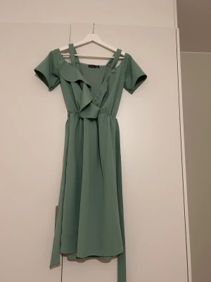 Grön omlottklänning från Boohoo - Snygg grön omlottklänning från Boohoo med korta ärmar och volangdetalj vid halsringningen. Klänningen har v-ringad front och ett knytband i midjan som ger en fin siluett. Perfekt för dig som gillar stilrena och trendiga plagg.