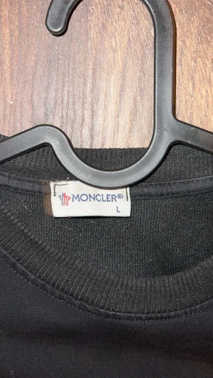 Svart sweatshirt från Moncler L - Svart sweatshirt från Moncler i storlek L med lång ärm och rund hals. Framsidan har ett stort broderat Moncler-märke i vitt och rött, samt en mindre logga på ärmen. Tröjan är gjord i mjuk bomull och har ribbade muddar vid hals, ärmslut och nederkant.