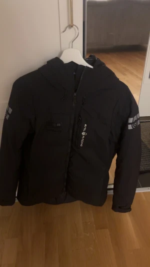 Svart Sail Racing dunjacka med huva - Svart dunjacka från Sail Racing, modell Antarctica Expedition. Jackan har huva, dragkedja framtill, logga på bröstet och ärmen samt flera praktiska fickor. Perfekt för kalla dagar och har en clean, sportig look.