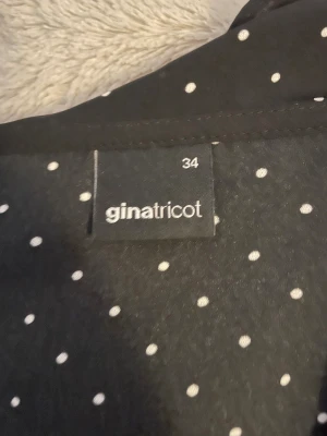 Svart prickig blus från Gina Tricot - Säljer en svart blus från Gina Tricot i storlek 34 med vita prickar och volangdetaljer längs halsringningen och ärmarna. Blusen har långärmade ärmar med knappar vid ärmslut och en omlott-look framtill. Perfekt för dig som gillar trendiga mönster och snygga detaljer.