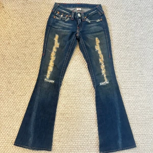 True Religion blå bootcut jeans - Snygga blå jeans från True Religion med bootcut passform och slitna detaljer framtill. Klassiska fickor med dekorativa sömmar och silverfärgade knappar. Jeansen har orange kontrastsömmar och grov tvättad look. Perfekt för dig som gillar street och retrostil.