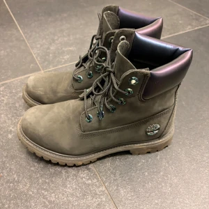 Svarta/grå Timberland boots med metallic-detaljer - Säljer ett par svarta Timberland boots med coola metallic-detaljer på öglor och logga. Skorna har en robust platt sula, snörning och en mjuk, skimrande lila kant upptill. Tillverkade i slitstark mocka, perfekta för dig som gillar street och outdoor-vibe.