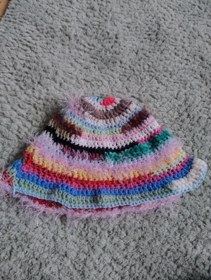 Färgglad virkad bucket hat - Unik bucket hat som är handvirkad i flera färger med mjuka och fluffiga detaljer. Hatten har ett randigt mönster i rosa, blått, gult, grönt, vitt och fler nyanser, samt glittriga inslag av fluffigt garn. Perfekt för dig som vill sticka ut med en kreativ och lekfull stil. Jag har virkat dne själv om någon undrar 🙃